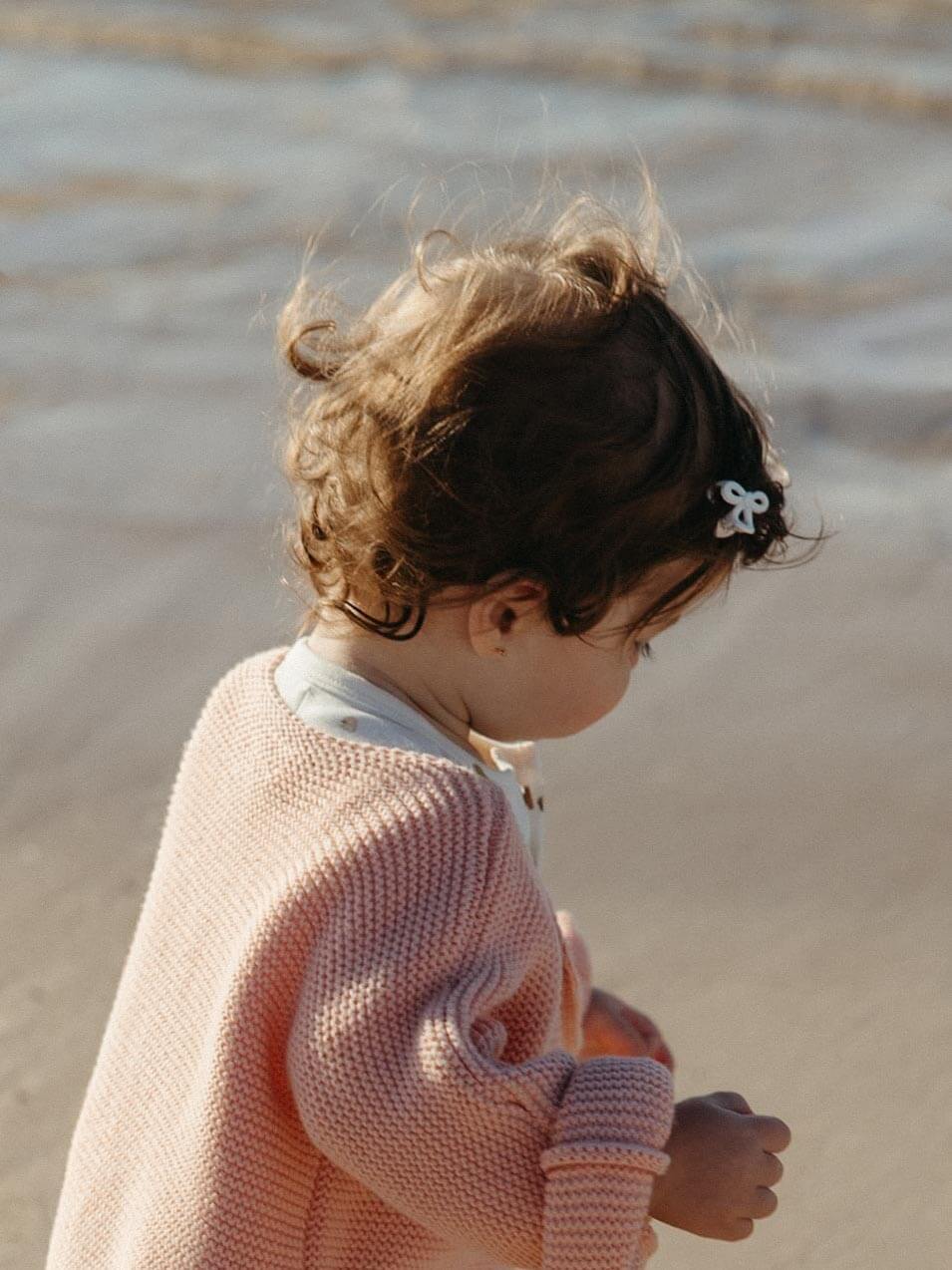 A toddler in profile wearing a soft pink knit cardigan and a small white bow hair clip, standing outdoors against a blurred sandy background.