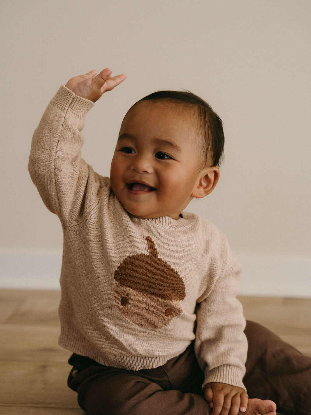 Baby in beige sweater with acorn design waving and smiling on wooden surface Baby Feature Print Jumper - Little Acorn Baby boy