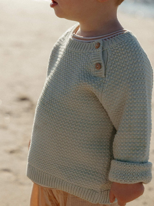 A close-up side view of a child wearing a textured knit sweater on a sandy beach. The sweater is a light oatmeal-beige color with a small button detail on the chest.