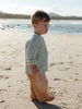 A toddler standing on a sandy beach wearing a textured pale sage green knit sweater and tan pants, with water and distant land in the background.