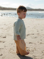 A toddler standing on a sandy beach wearing a textured pale sage green knit sweater and tan pants, with water and distant land in the background.