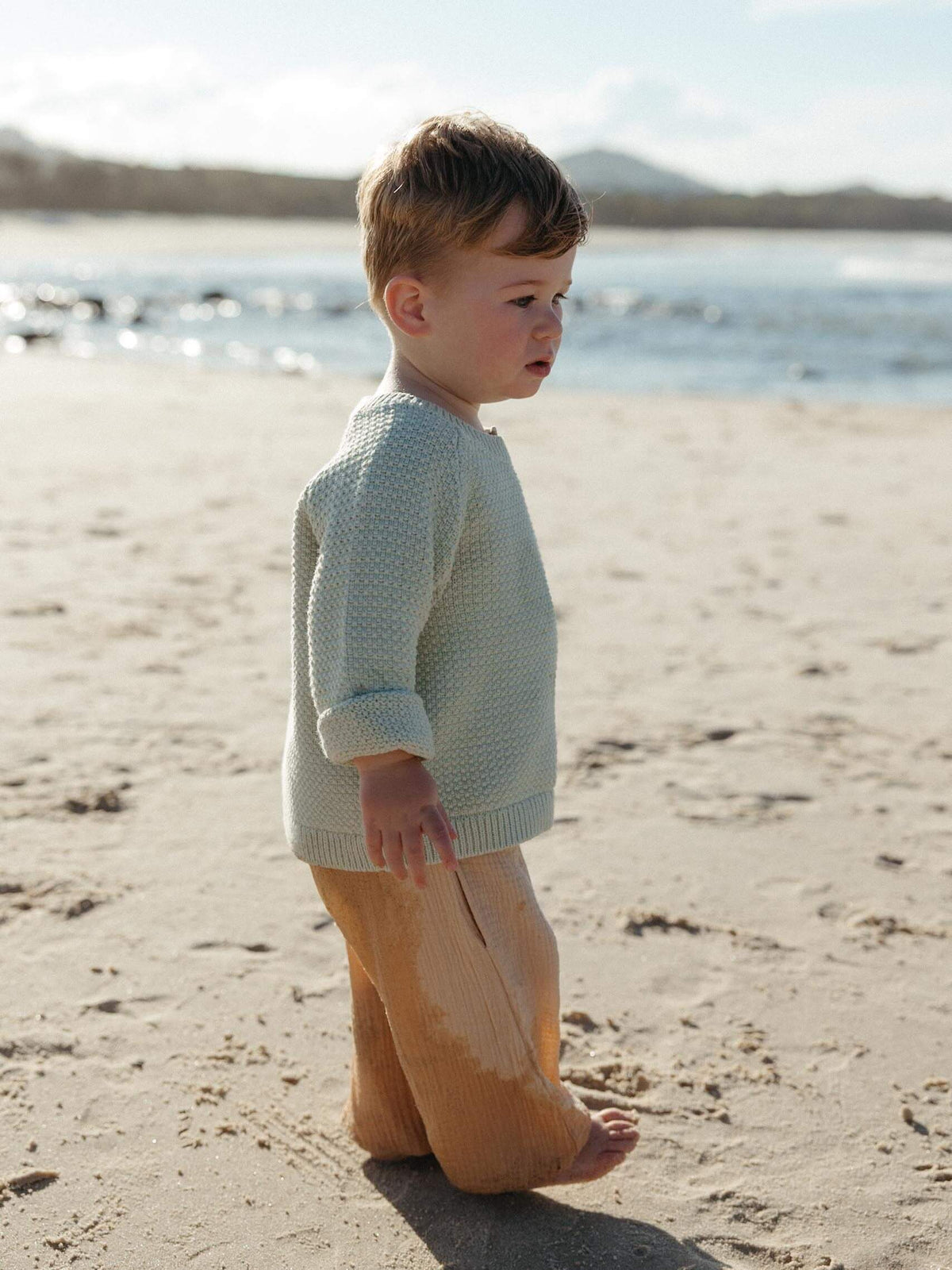 A toddler standing on a sandy beach wearing a textured pale sage green knit sweater and tan pants, with water and distant land in the background.