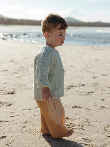 A toddler standing on a sandy beach wearing a textured pale sage green knit sweater and tan pants, with water and distant land in the background.