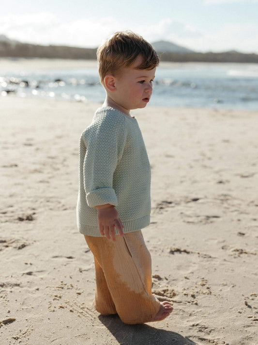 A toddler standing on a sandy beach wearing a textured pale sage green knit sweater and tan pants, with water and distant land in the background.
