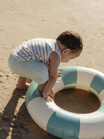 baby boy playing with a beach float on the sand wearing a light green stripe shorts and a striped sleeveless tee