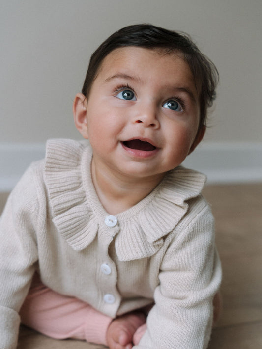 Baby wearing cream knit cardigan with ruffle collar and white buttons Baby Ruffle Cardigan - Ecru Baby girl