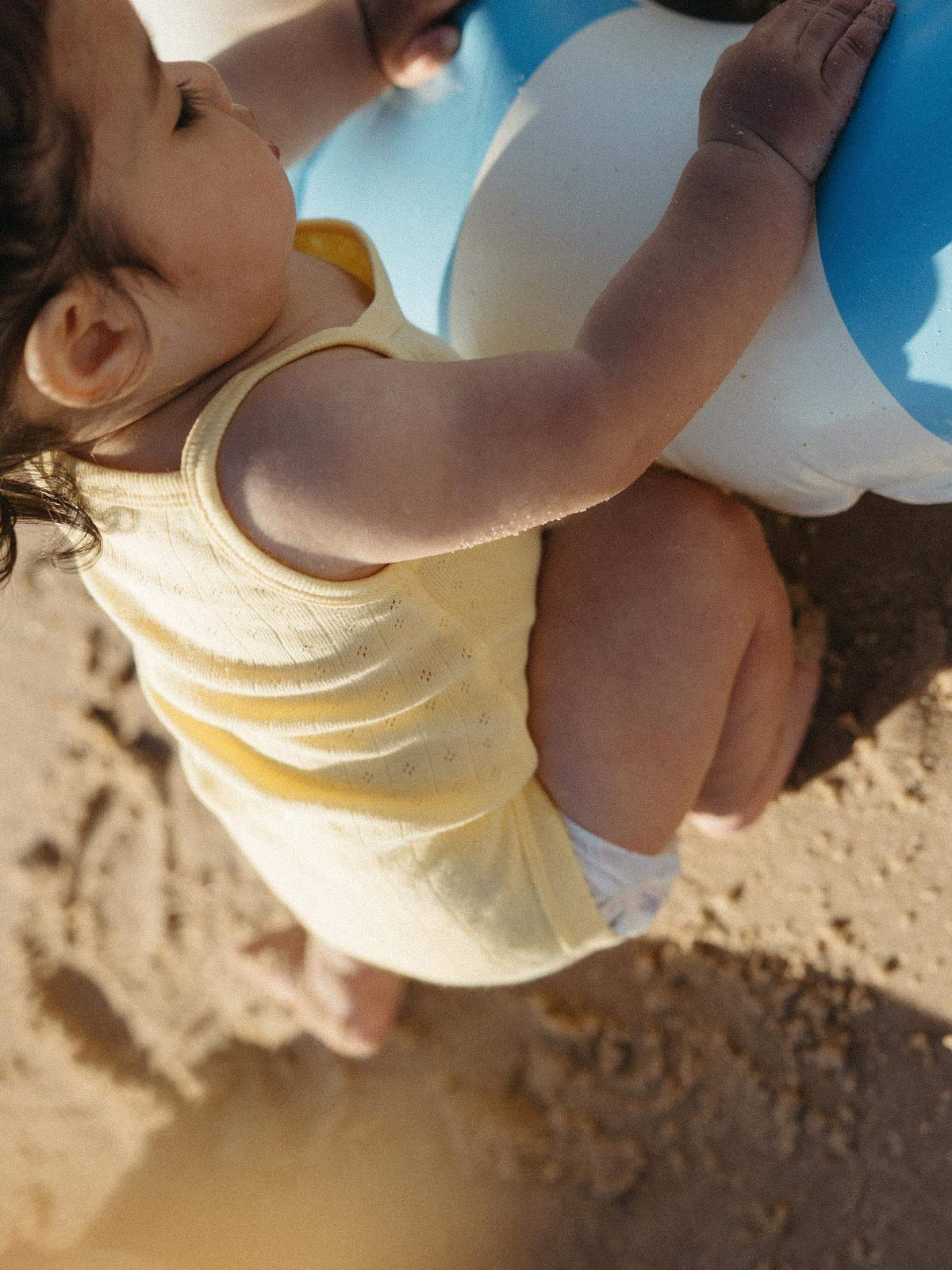 A close-up lifestyle shot of a baby wearing a pale yellow sleeveless tank top and a white diaper, sitting on sand and holding onto a blue and white beach ball.