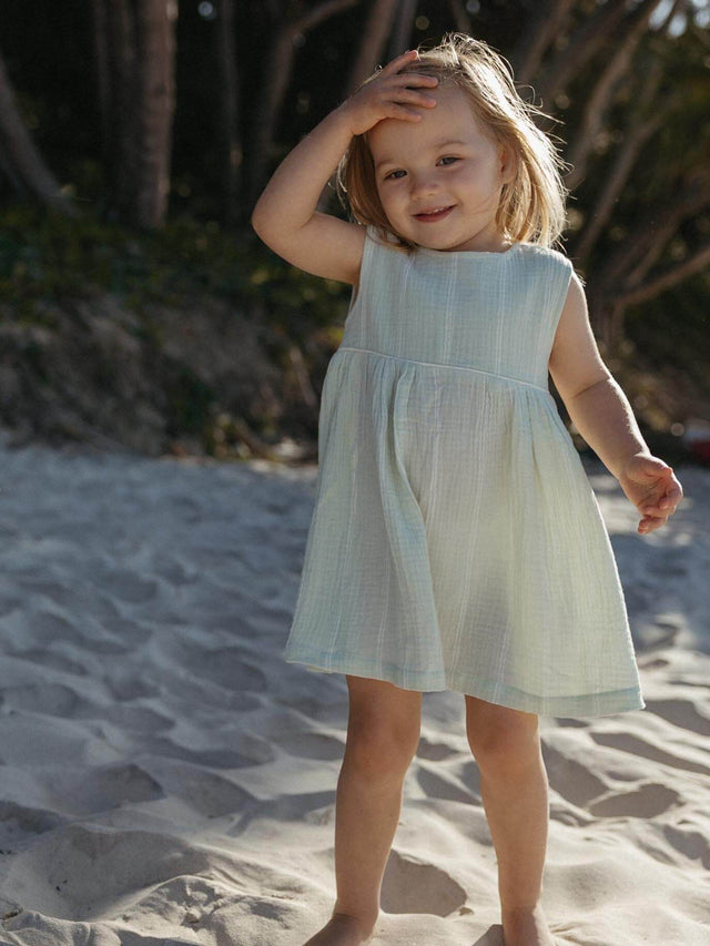 A young child wearing a sleeveless light blue textured dress, standing on a sandy beach with trees in the background.