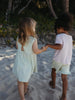 Two children holding hands walking on a sandy beach, viewed from behind. The child on the left wears a pale mint green sleeveless dress, and the child on the right wears a light pink t-shirt and pale green shorts.