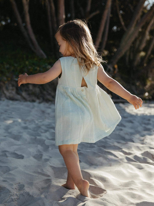 A young girl with long light brown hair stands on a sandy beach wearing a sleeveless, light blue-green textured dress with a back cutout. Trees are visible in the background.
