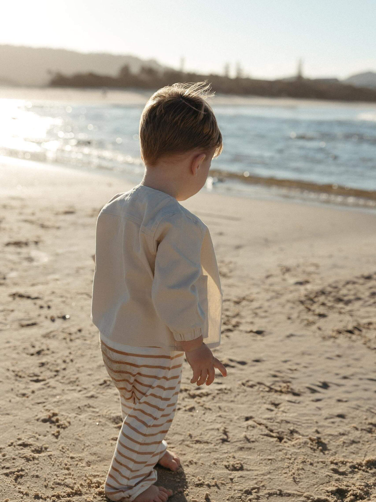 A toddler standing on a sandy beach at sunset, wearing a cream long-sleeve top and beige-and-white striped pants, looking toward the water.