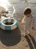 A toddler stands barefoot in shallow water on a sandy beach next to a striped inflatable pool float. The child is wearing a cream-colored zip-up jacket over a beige and white striped one-piece outfit.