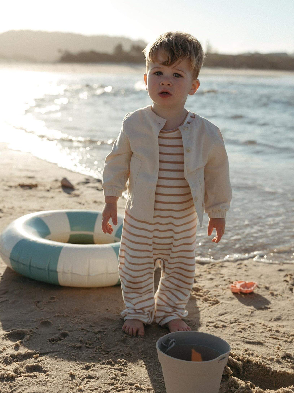 A toddler stands on a sandy beach wearing a cream-colored jacket over a cream and orange striped one-piece. A blue and white inflatable ring and a small bucket are on the sand nearby, with the ocean and shoreline in the background.