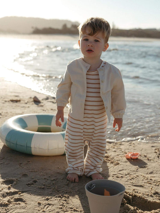 A toddler stands on a sandy beach wearing a cream-colored jacket over a cream and orange striped one-piece. A blue and white inflatable ring and a small bucket are on the sand nearby, with the ocean and shoreline in the background.