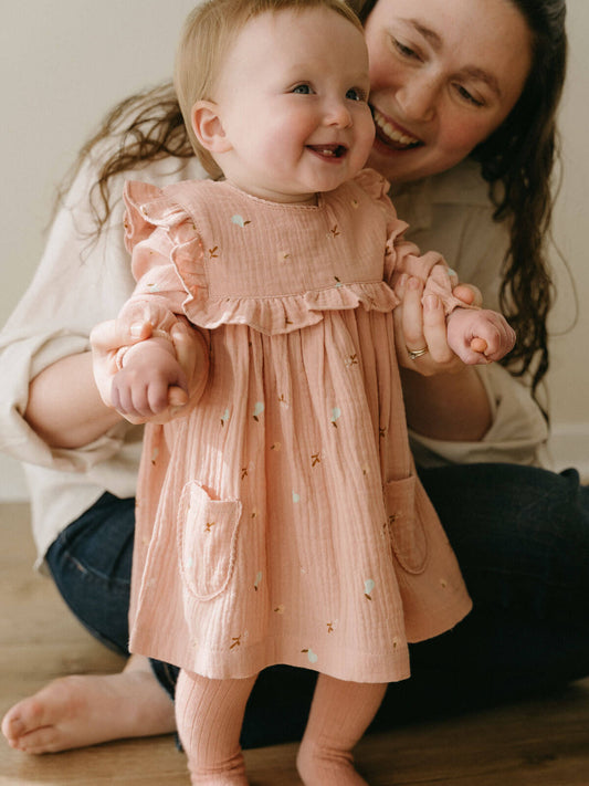 Woman holding a baby wearing a pink dress with ruffled sleeves and a pocket. Baby Woven Dress - Autumn Harvest Rosehip Baby girl