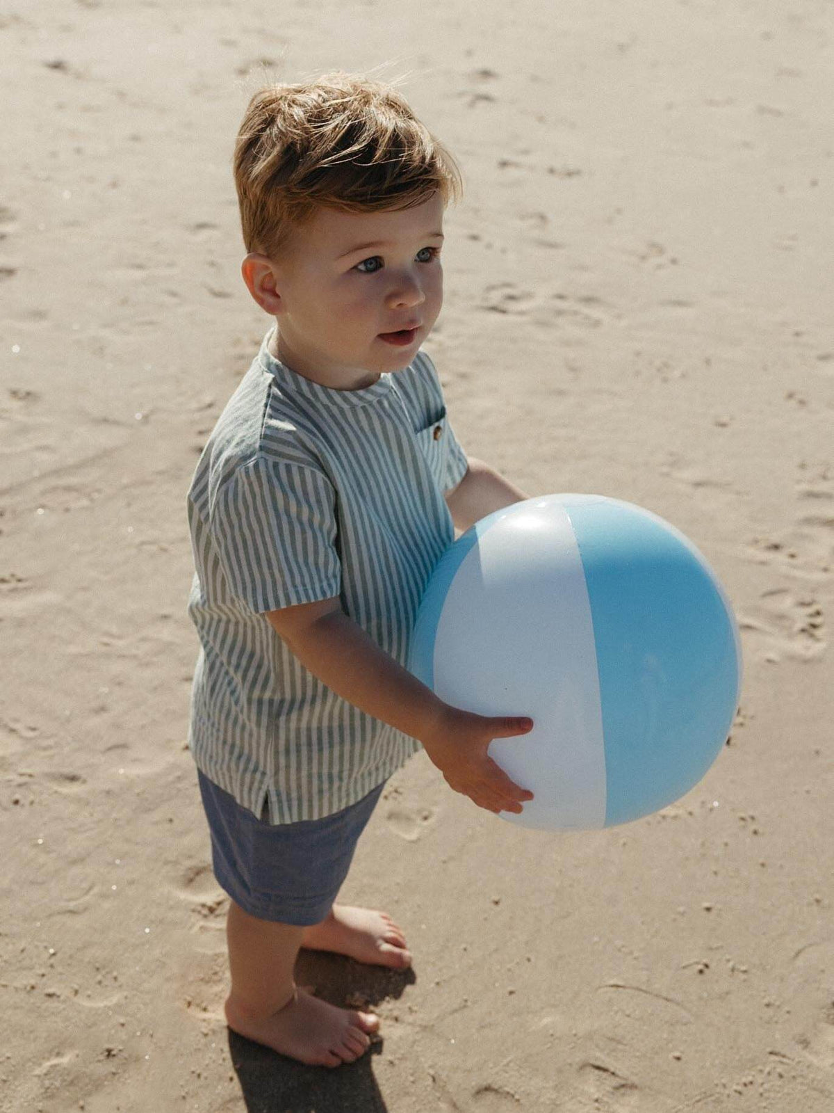 A young boy standing on a sandy beach holding a light blue and white beach ball. He is wearing a light blue and white vertically striped short-sleeve shirt and blue shorts.