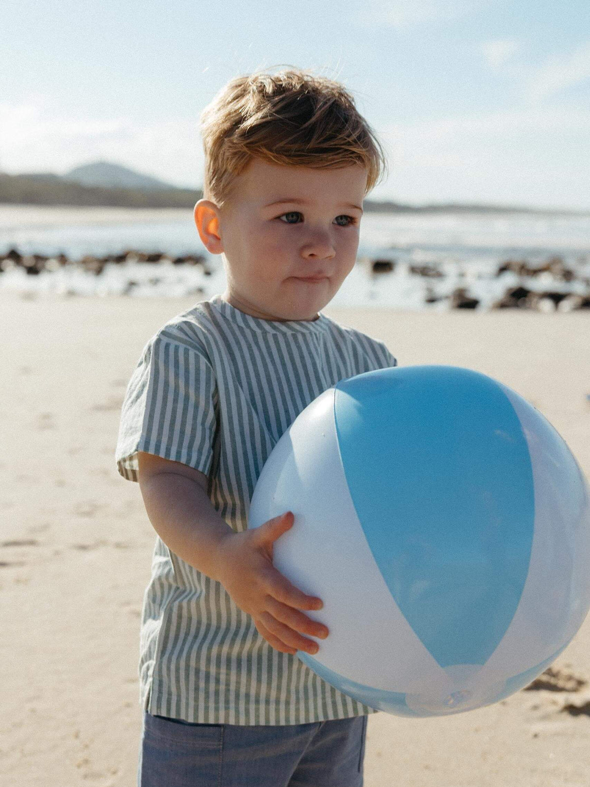 A young boy standing on a sandy beach holding a large blue and white beach ball. He is wearing a light blue and white vertically striped short-sleeve t-shirt and blue jeans.
