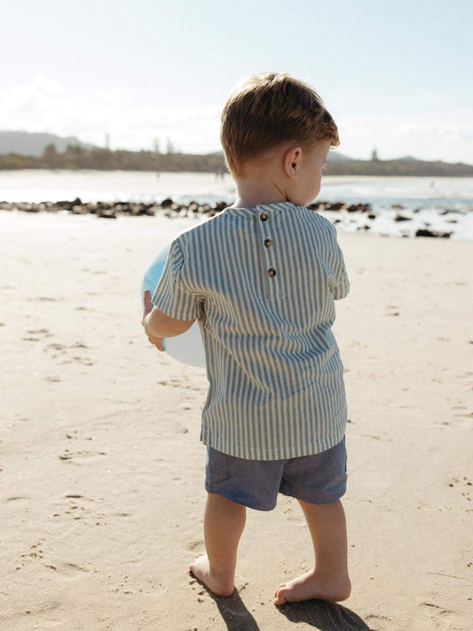 A toddler standing on a sandy beach, viewed from behind, wearing a light blue and white vertically striped short-sleeve shirt with three brown buttons at the back and blue shorts.
