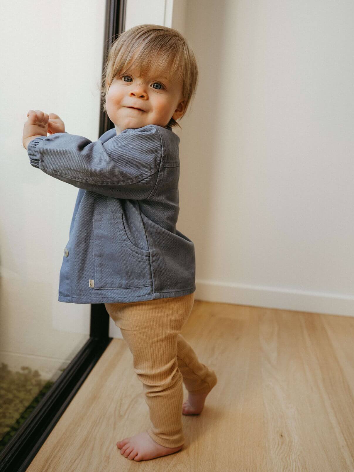 Child wearing a denim jacket and beige pants standing near a window.