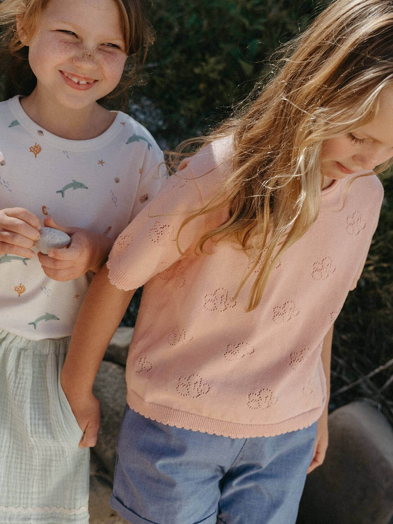 Two children standing outdoors, one in a light pink sweater and the other in a white shirt with patterns.