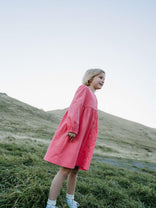 Girl wearing a pink dress standing on a grassy hill with a clear sky. Button Through Dress - Cloudberry Girls