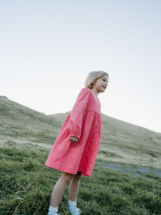 Girl wearing a pink dress standing on a grassy hill with a clear sky. Button Through Dress - Cloudberry Girls