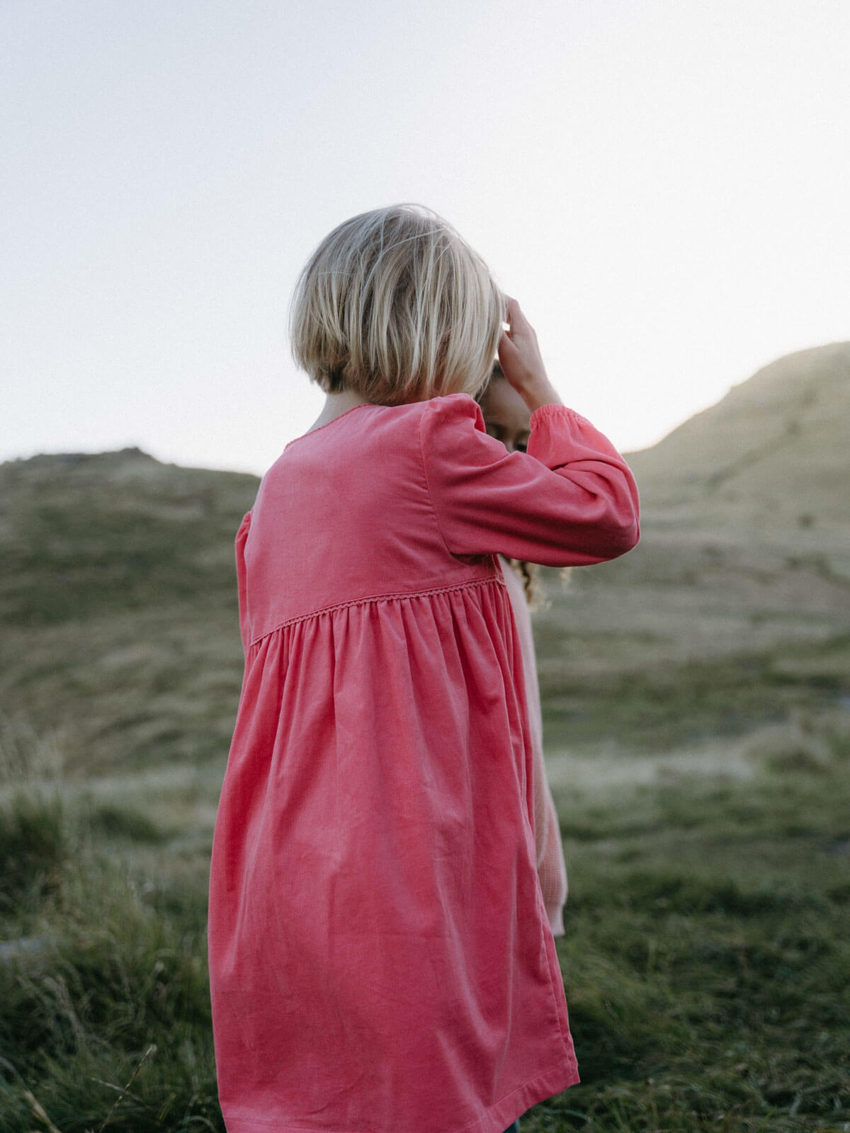 Girl wearing a pink dress standing in a grassy field with mountains in the background Button Through Dress - Cloudberry Girls
