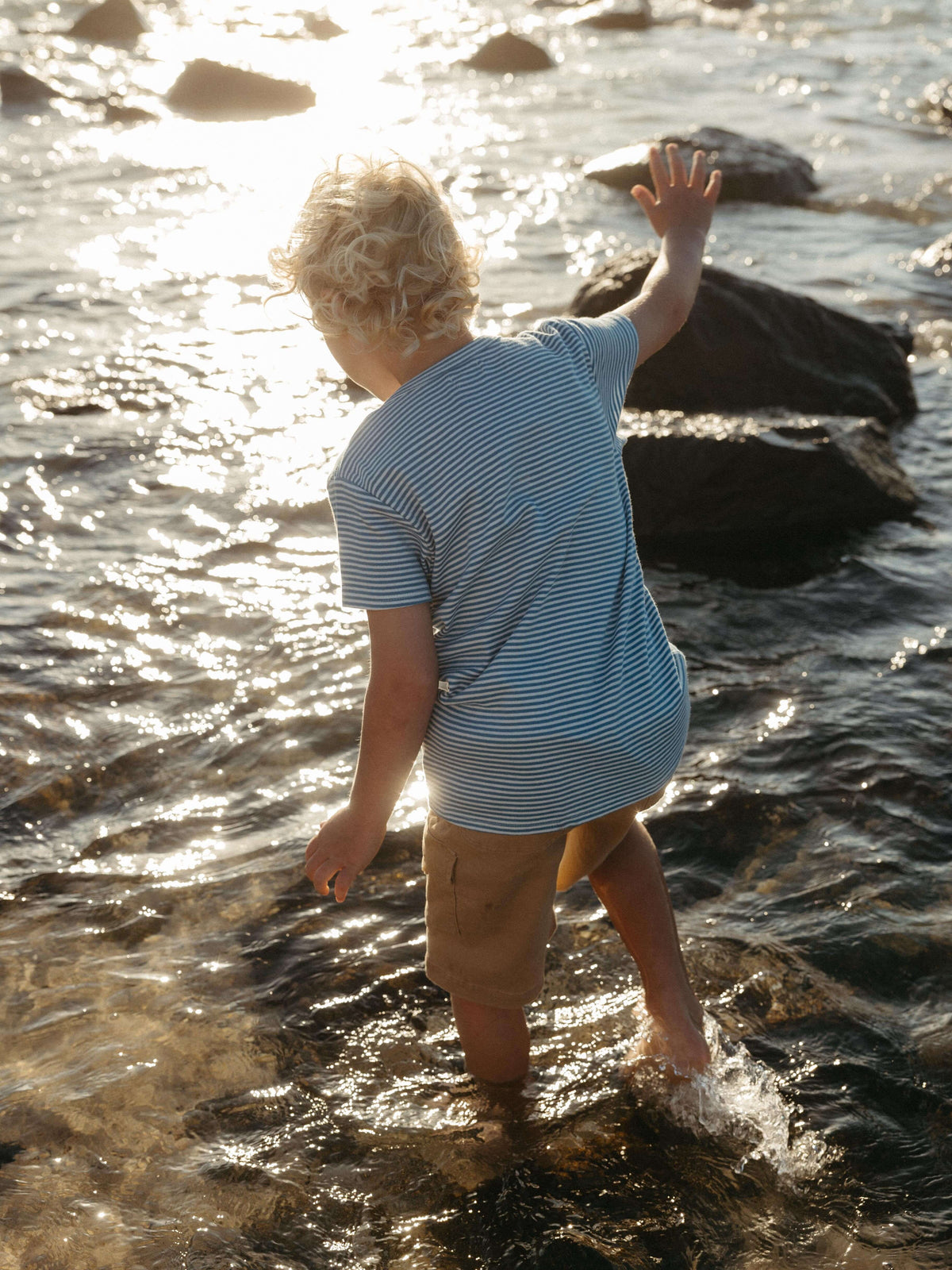 A young child with blonde hair stands in shallow water at a beach or lake, seen from behind, wearing a light blue and white striped t-shirt and tan shorts, with rocks and sparkling sunlight in the water.