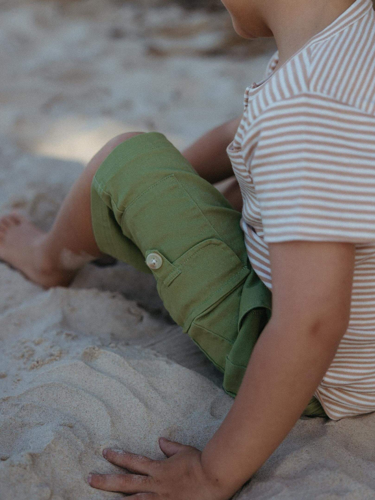 A child sitting on sandy ground wearing olive green cargo shorts and a cream and brown striped t-shirt.