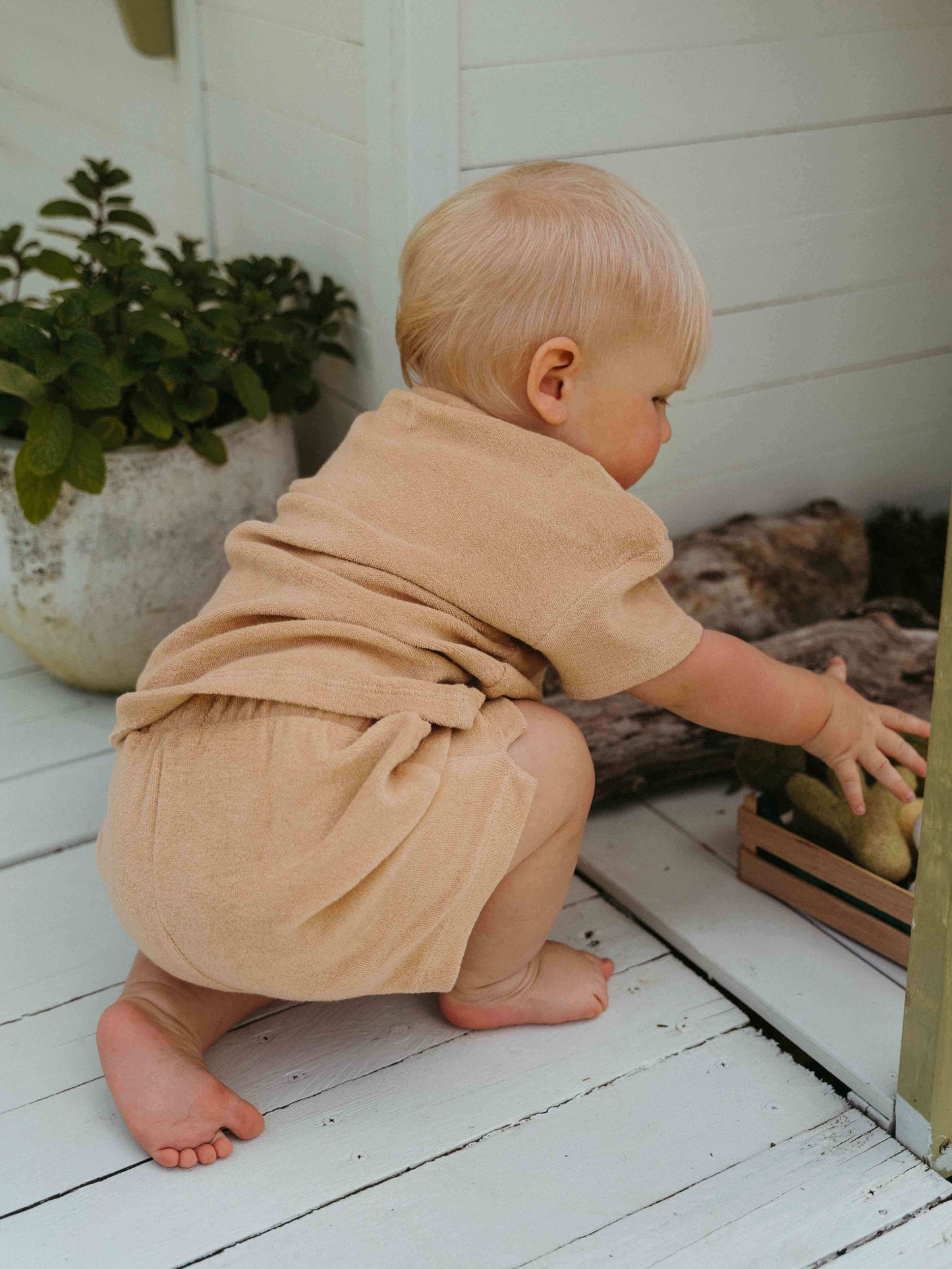 Child in a beige outfit playing with a toy on a white wooden floor. Carter Towelling Shorts - Sand Baby boy