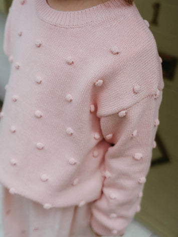 Pink knitted sweater with pom-pom details on a blurred background Clara Jumper - Candy Floss Girls