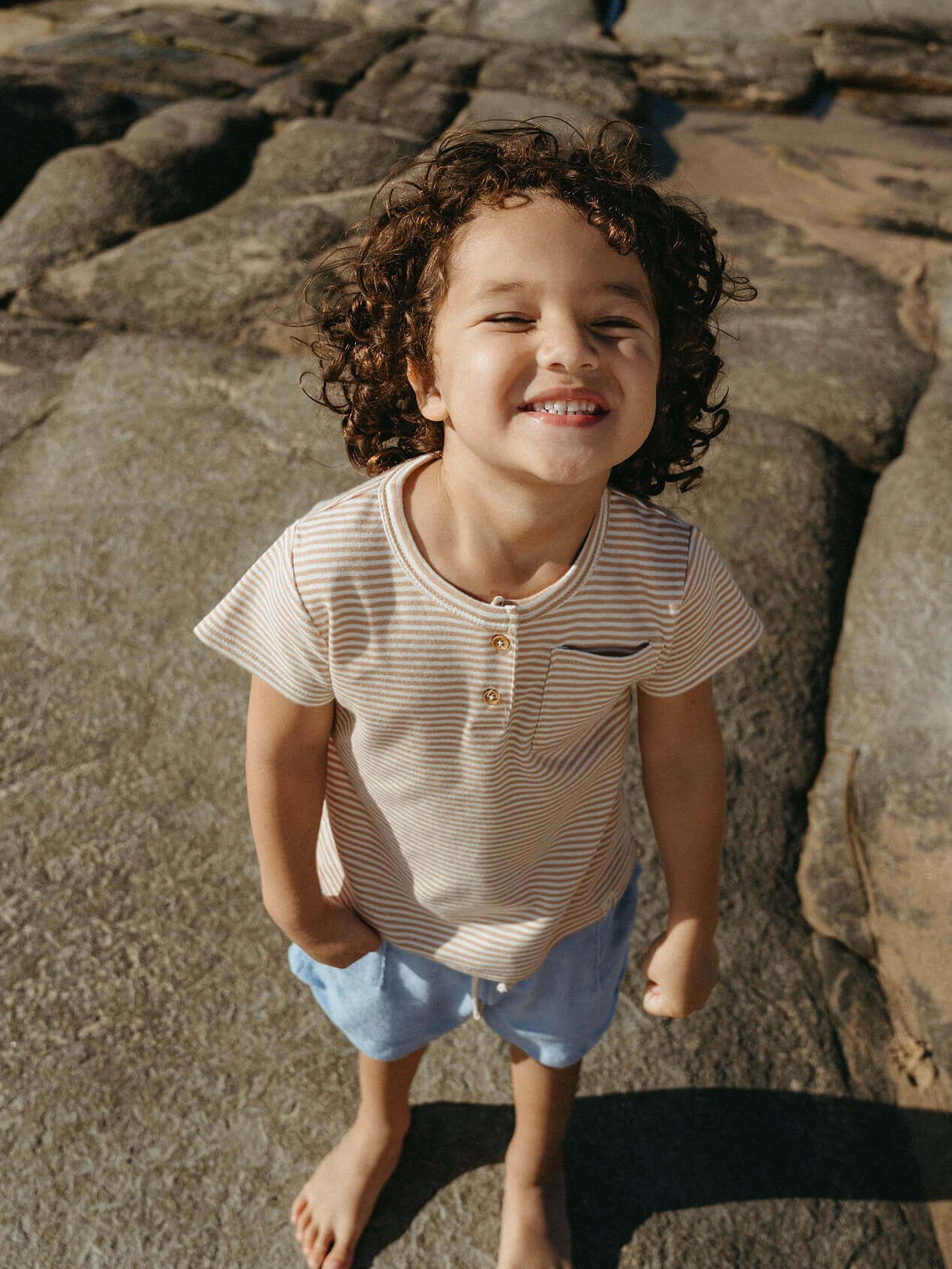 Child standing on rocks wearing a striped shirt and shorts, smiling.
