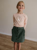 Young girl wearing a pink shirt with a graphic and a green skirt standing on a wooden floor.