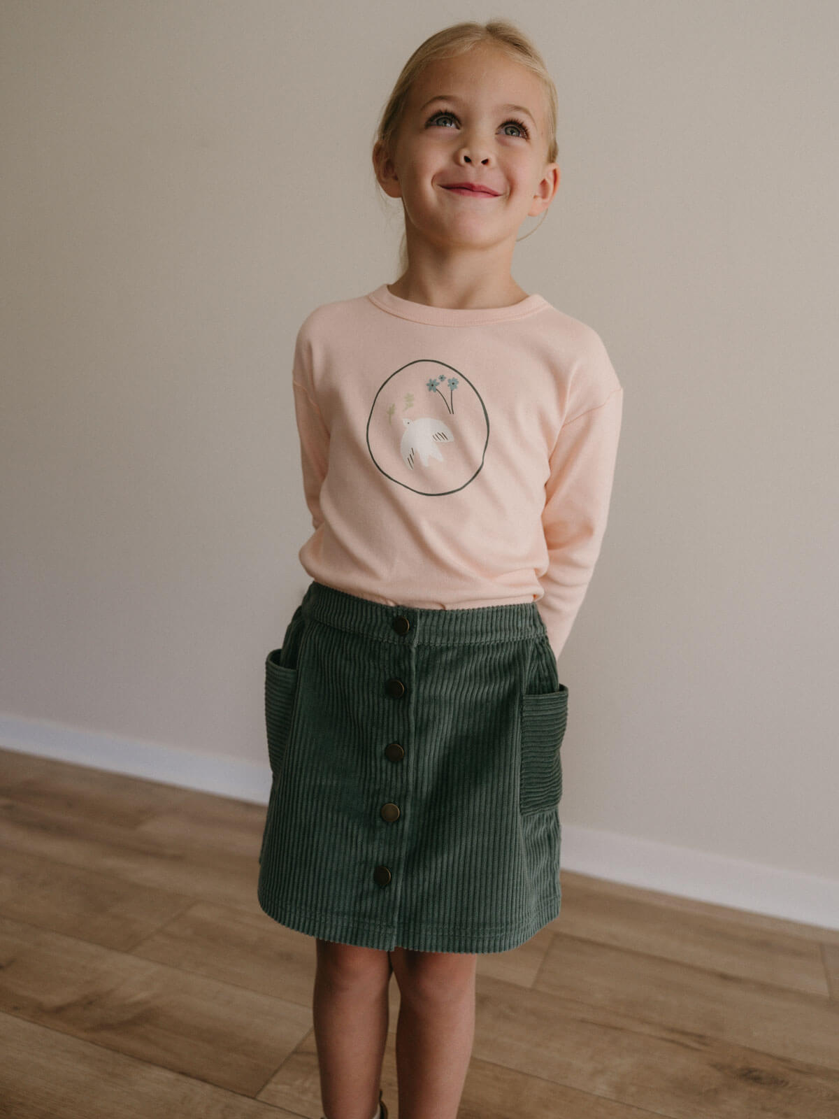 Young girl wearing a pink shirt with a graphic and a green skirt standing on a wooden floor.