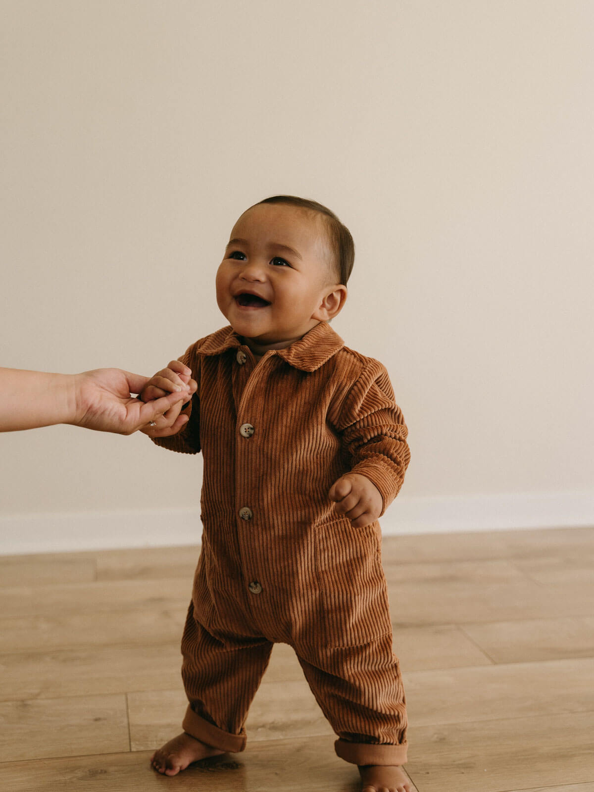 Baby in a brown corduroy outfit standing on a wooden floor with a blurred background Corduroy Jumpsuit - Russet Baby boy