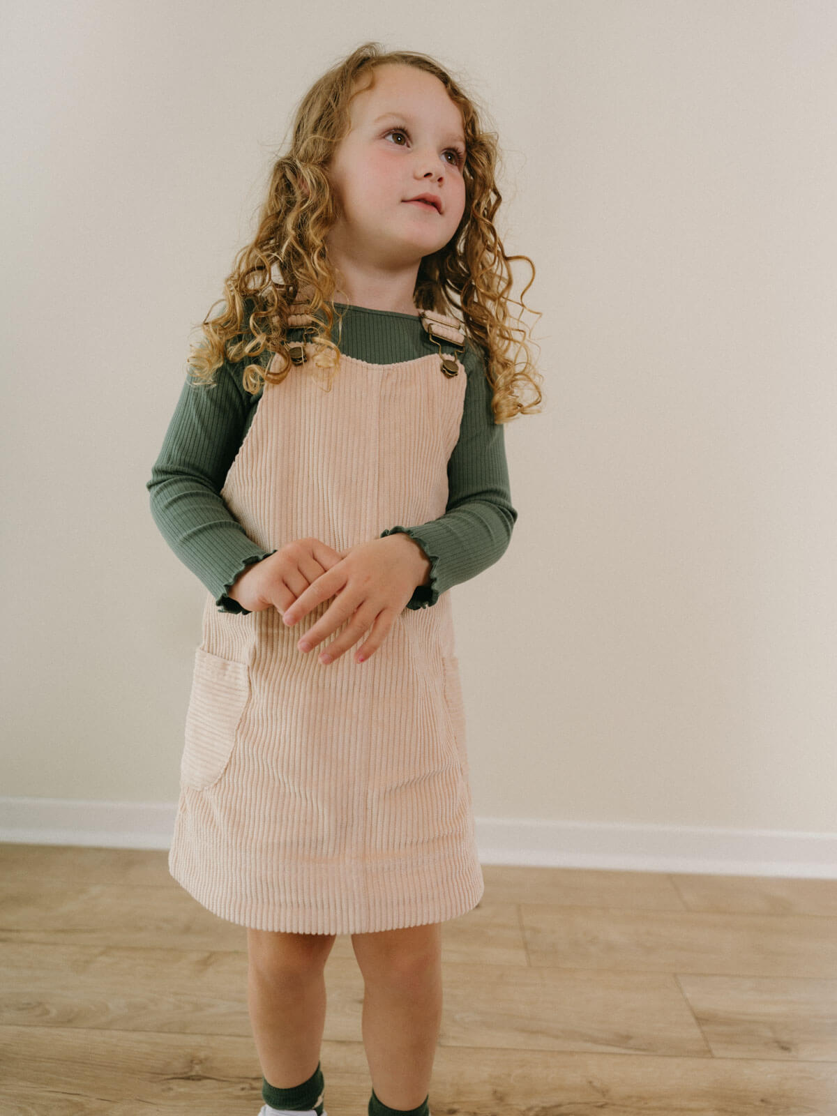 Young girl wearing a green long-sleeve shirt and beige pinafore dress standing on a wooden floor. Corduroy Pinafore Dress - Clay Girls