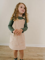 Young girl wearing a green long-sleeve shirt and beige pinafore dress standing on a wooden floor. Corduroy Pinafore Dress - Clay Girls