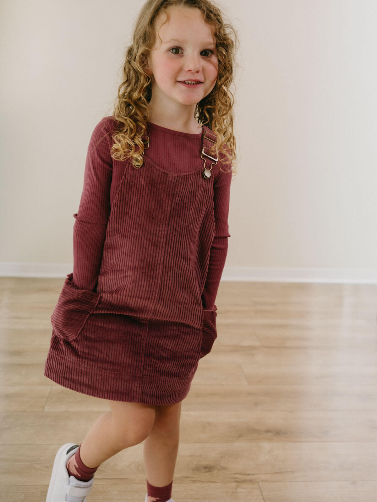 Young girl wearing a maroon corduroy dress standing on a wooden floor.