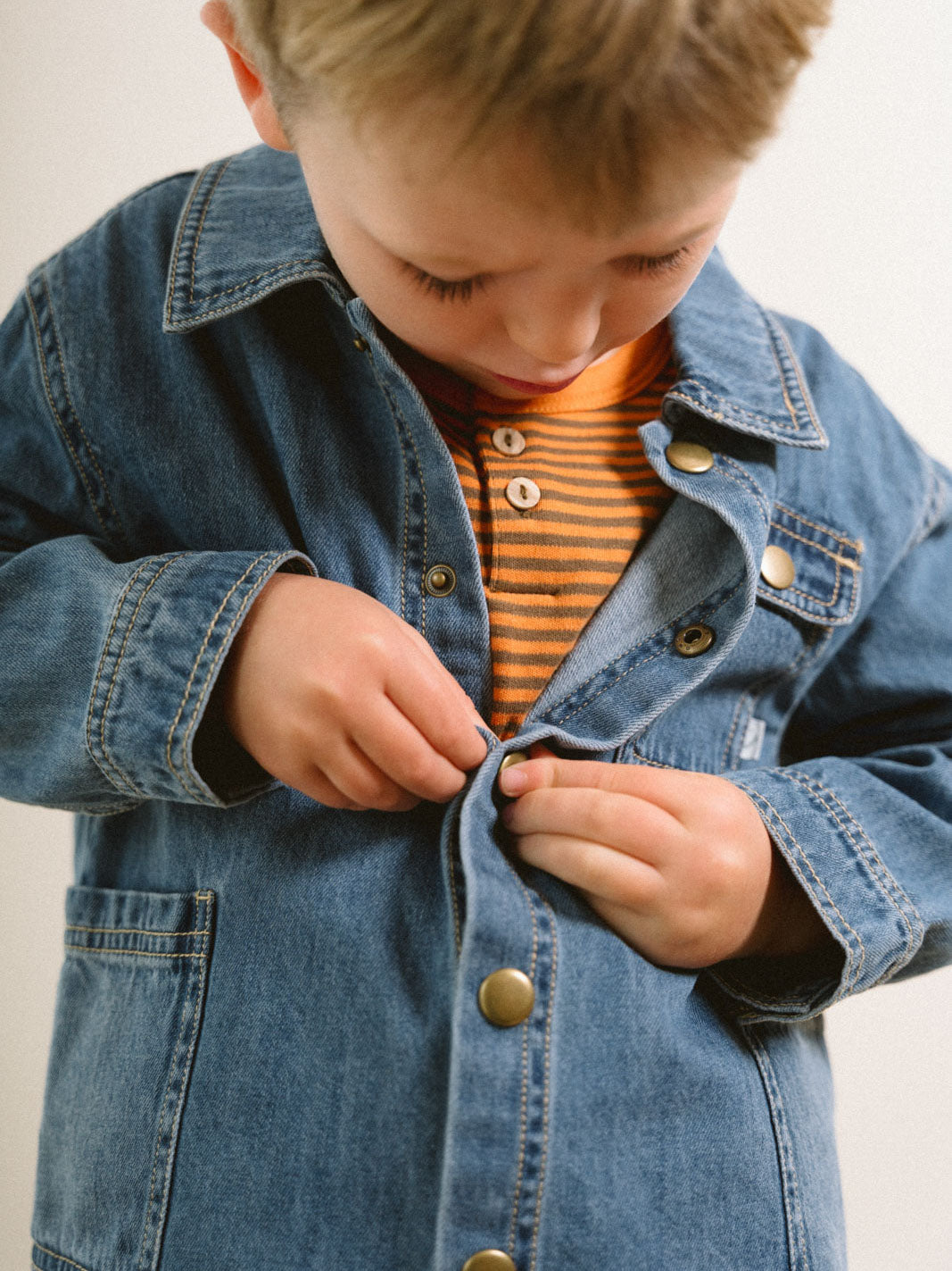 Young child buttoning up a blue denim jacket over a striped shirt Denim Jacket - Blue Denim Girls and Boys