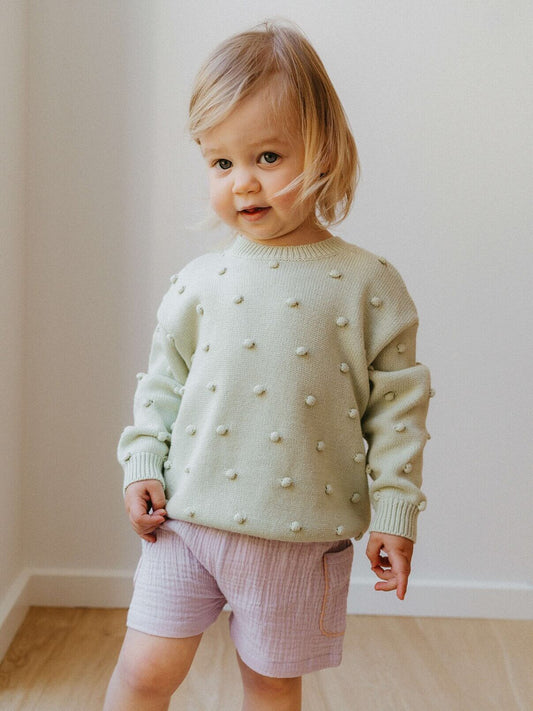 Child wearing a light green sweater with white polka dots and pink shorts indoors.