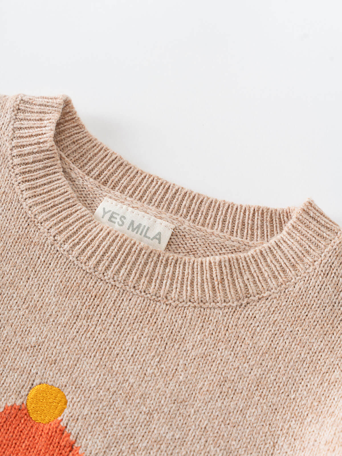 Close-up of a beige sweater with a floral design and 'YES MILA' label on a white background