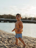 A toddler walking on a sandy beach near the water, wearing a peach-orange long-sleeve sweatshirt with a blue graphic and light blue denim shorts.