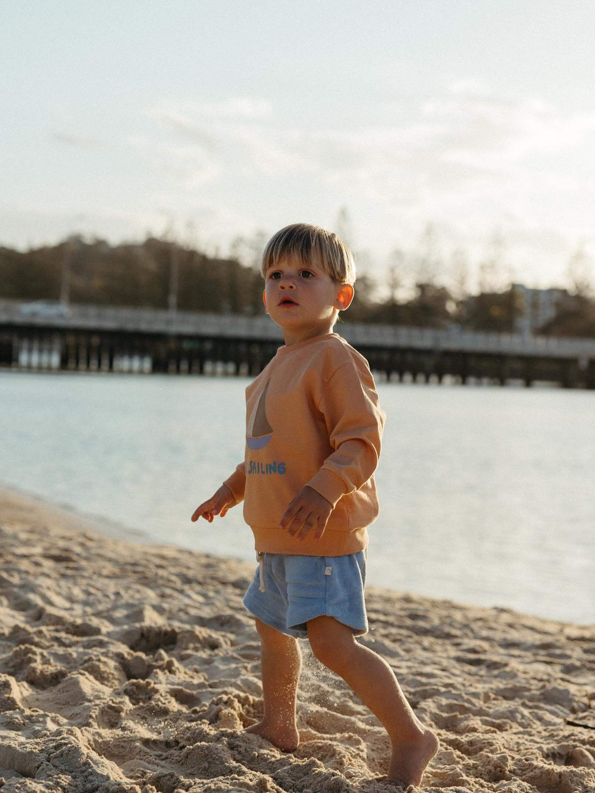 A toddler walking on a sandy beach near the water, wearing a peach-orange long-sleeve sweatshirt with a blue graphic and light blue denim shorts.