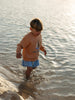 little boy walking on the water at the beach wearing an orange coloured sweatshirt and a blue shorts