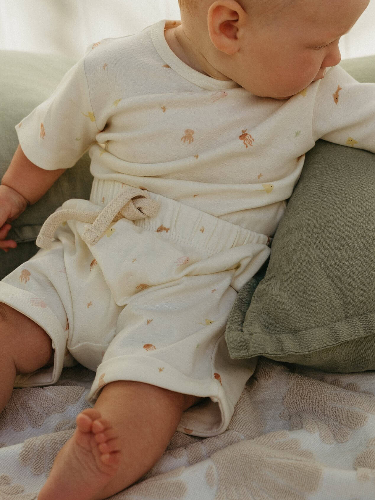 Baby in a white outfit with small patterns sitting on a green cushion.