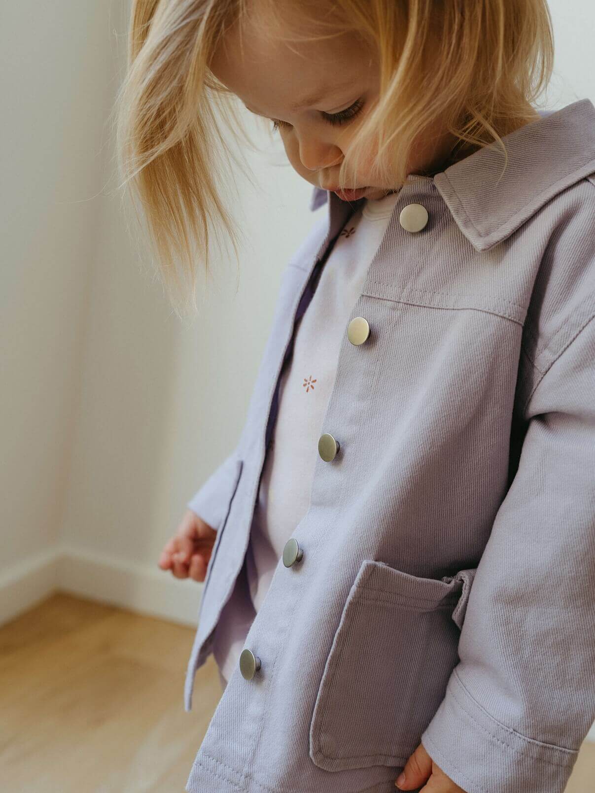 Child wearing a light purple coat sitting on a wooden floor.