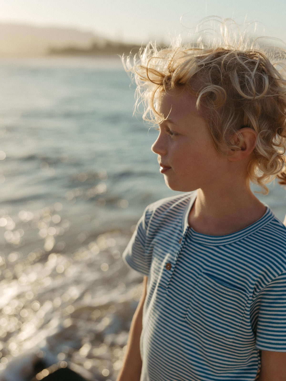 A young child with curly blond hair stands by the water at sunset, wearing a blue and white striped short-sleeve shirt.