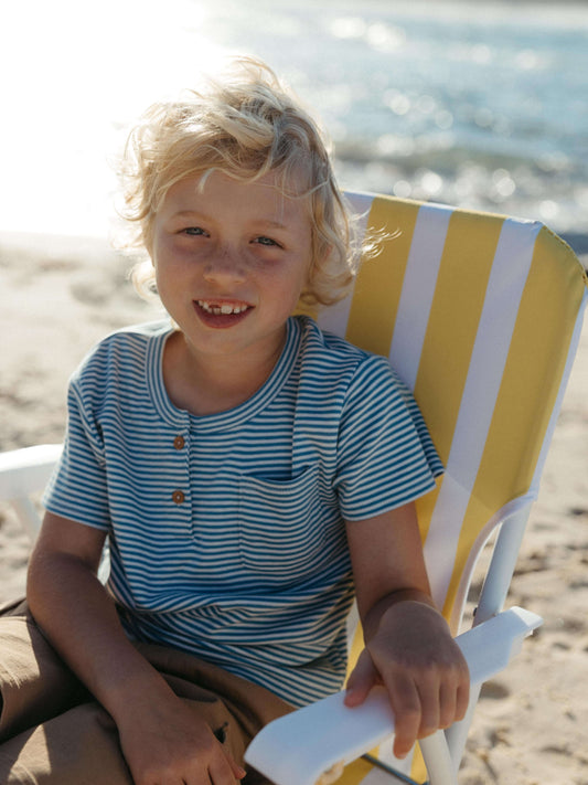 A young blonde boy smiling while sitting on a yellow and white striped beach chair on a sandy beach, wearing a blue and white striped henley shirt.