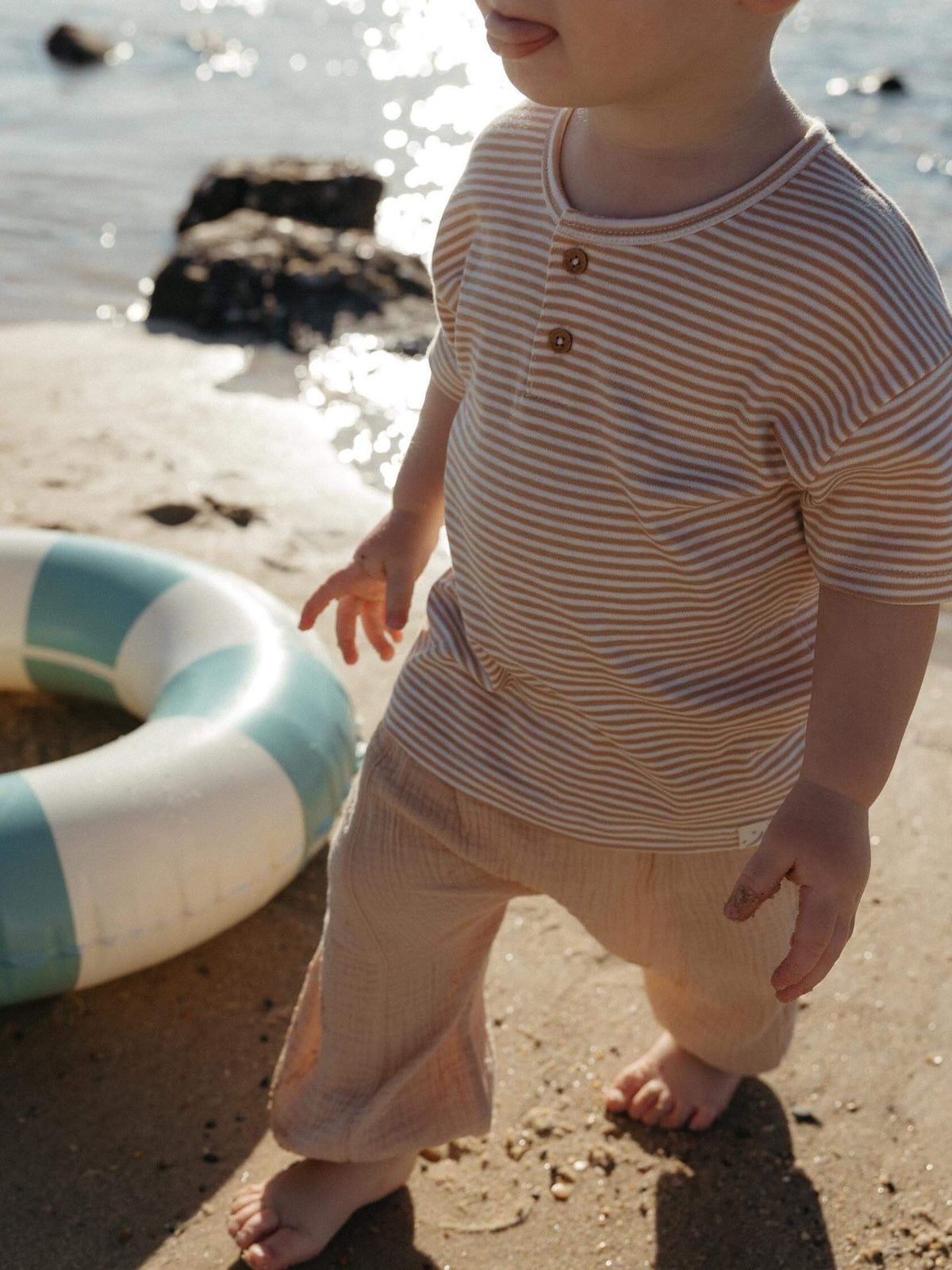 Toddler walking on the beach wearing a caramel colour pants and a striped baby tee