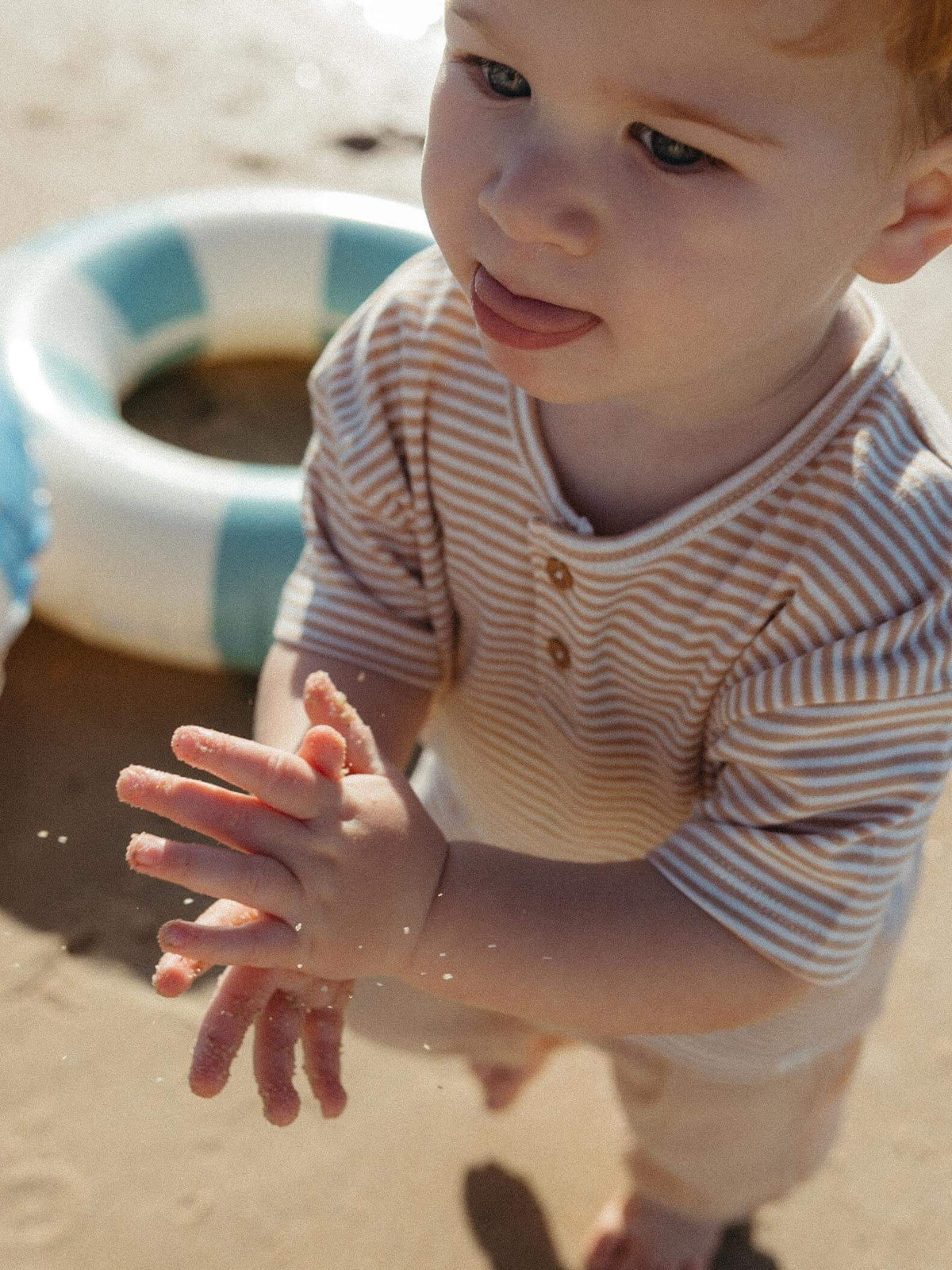 toddler boy playing with sand in his hands wearing a caramel striped tee